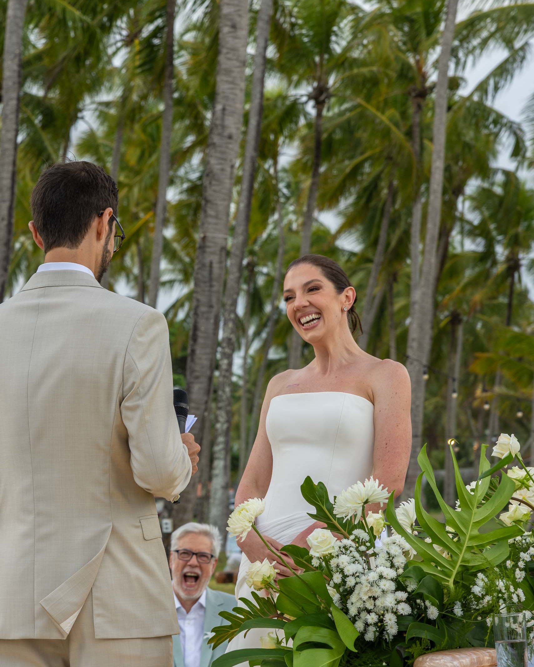 Bride smiling as she listens to her partner during an outdoor beach wedding ceremony surrounded by tropical palm trees.